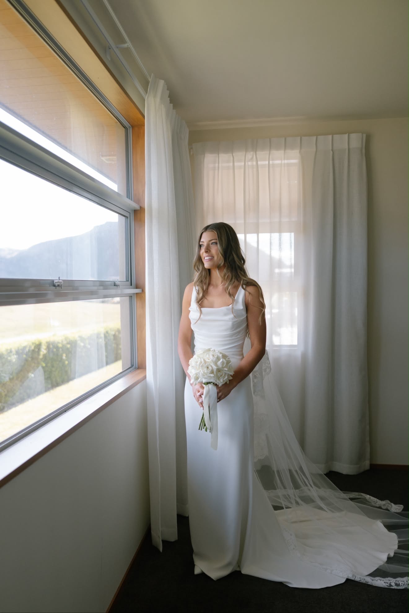 Bride with a soft bridal spray tan standing by the window with her bouquet